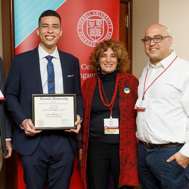 		Conor Hodges in suit and tie, smiling and holding his award plaque, flanked by other alumni.
	