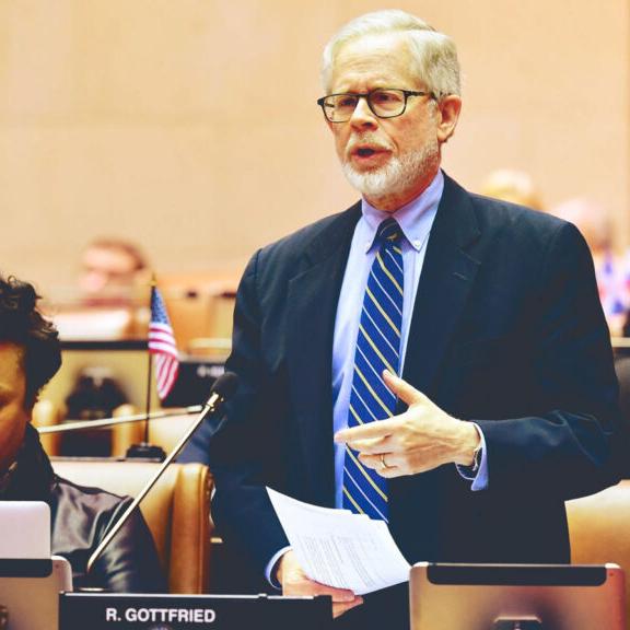 		Person wearing a suit standing behind a desk, speaking seriously
	