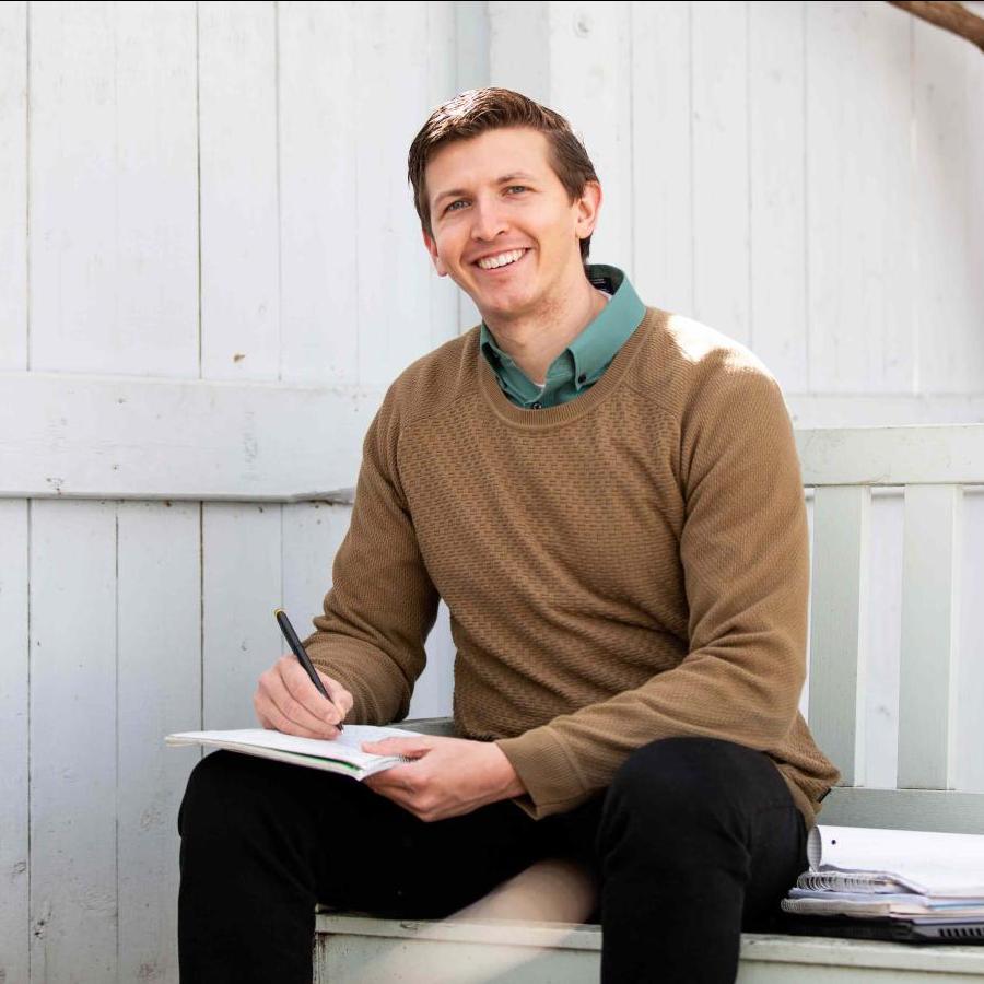 		J.J. Zanazzi sitting on a white wooden bench underneath a leafless tree, writing in a notebook; smiling.
	