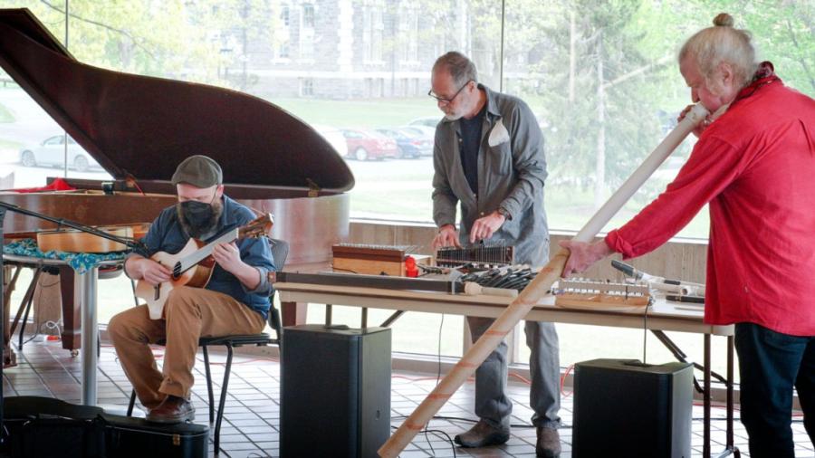 Three musicians: one playing a strangely shaped guitar, one playing a long tube half white plastic half cardboard, and one playing a metal sort of keyboard.