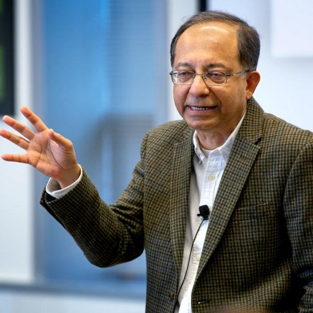 Kaushik Basu wearing a tweed jacket with hand upraised as he delivers a talk.