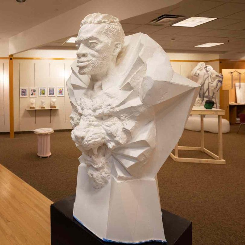 An African man's head with a ray-like collar above the face of a roaring lion with other artwork from the Sculpture Shoppe exhibition in the background.