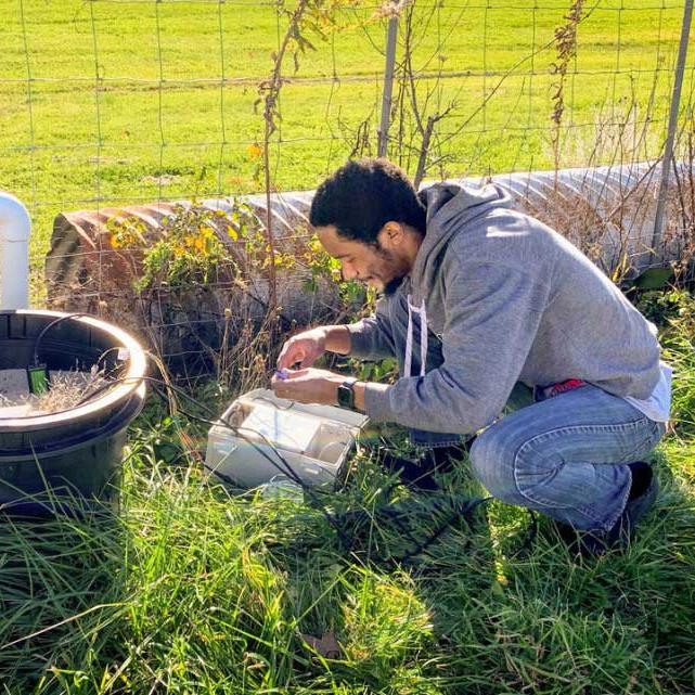 Person crouching in a field, tinkering with a device near a fence