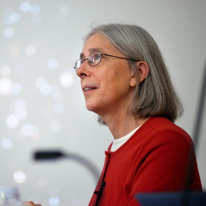 Martha Haynes with glasses, shoulder-length gray hair in a red top, with blurred stars on screen behind her