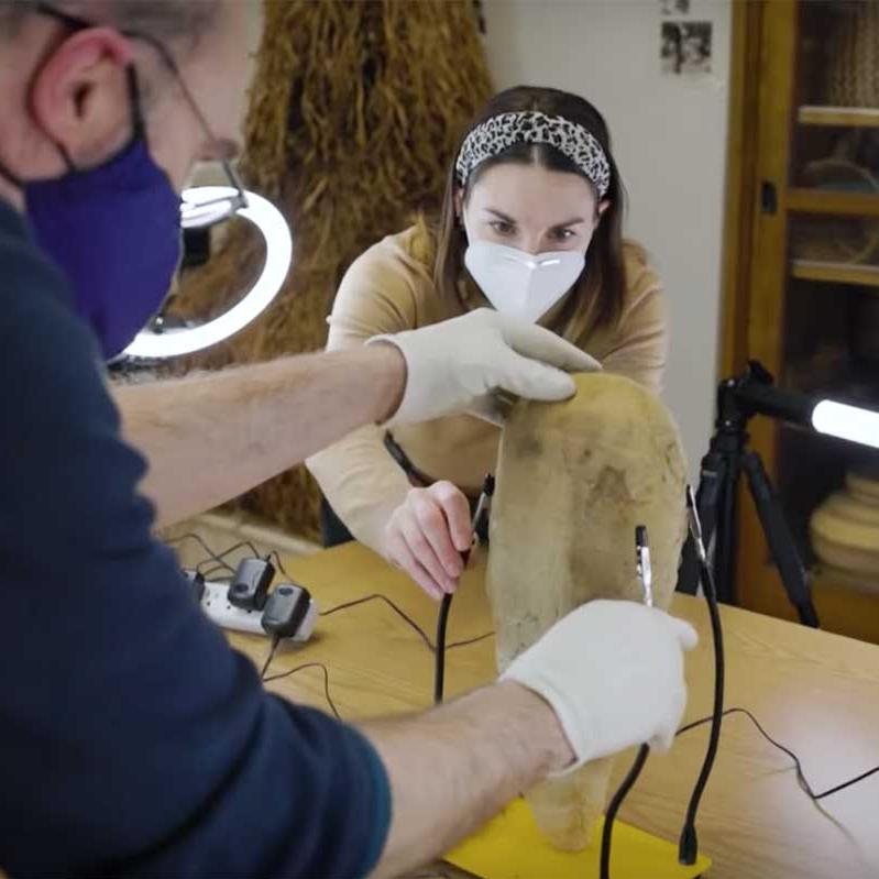 Two people wearing gloves work with football-sized museum object