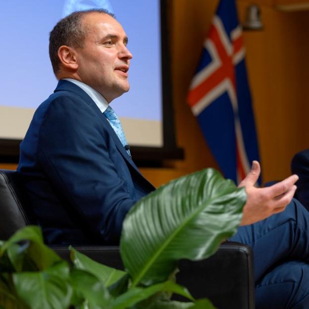 Two people wearing suits speak, seated on a stage among plants