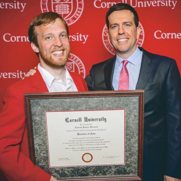 Two people stand in front of a red backgroun, holding a framed diploma