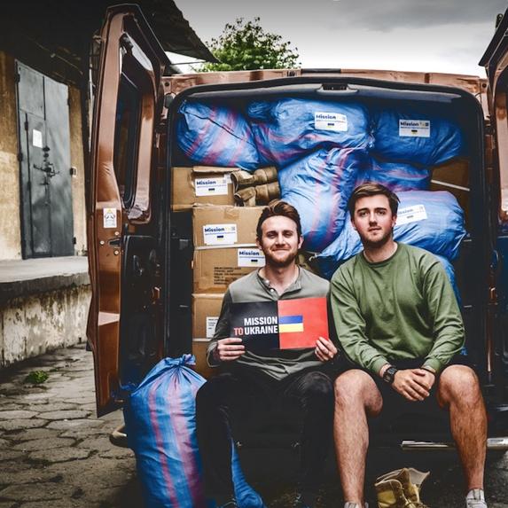 Two people sitting in the back of a van with doors open, showing boxes and bags