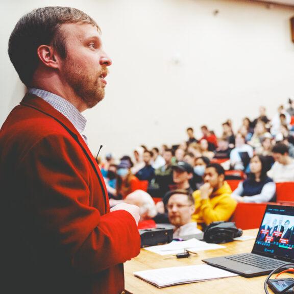 Person in red jacket speaks to a full auditorium