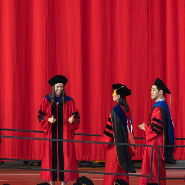Several people in long red robes and black caps walk in a line against a red background; one turns to give a thumbs up