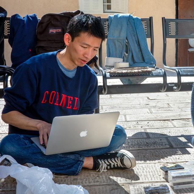 Two people sit on the ground; one types on a laptop, the other holds a compact disc