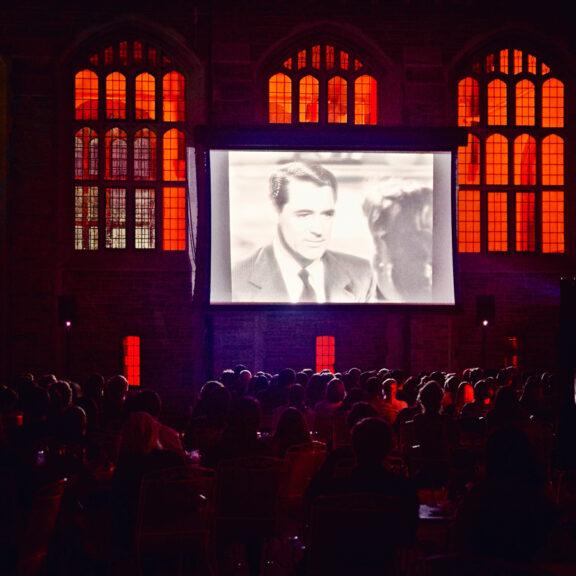 Movie screen outdoors, showing a black and white still of Jimmy Stewart, with red-lit windows behind it. 