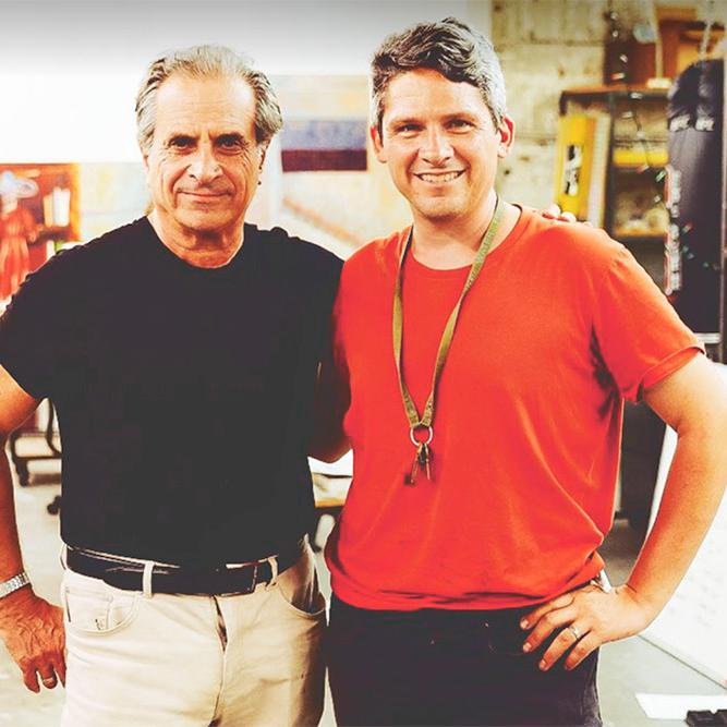 		Two people stand together in a studio with paints and paintings
	