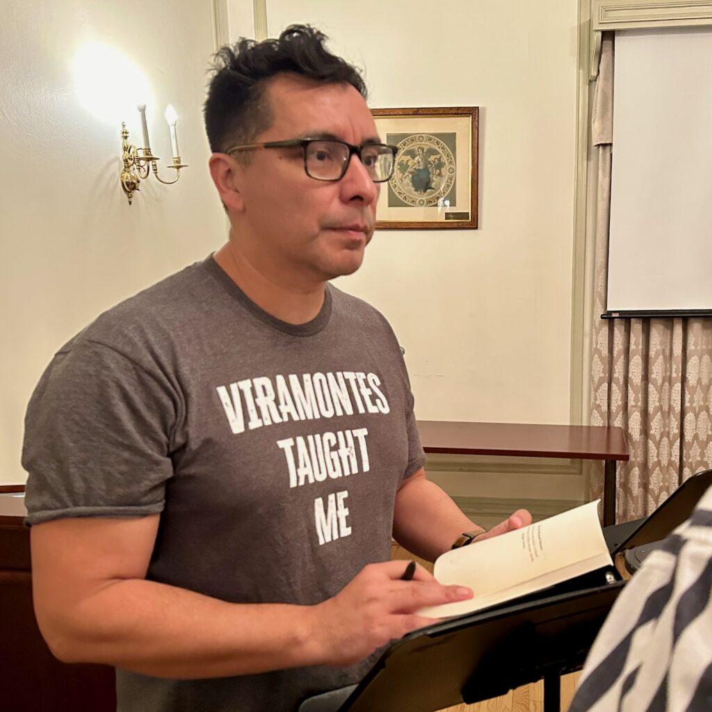 		Person standing at a podium, holding a book, with a serious expression
	