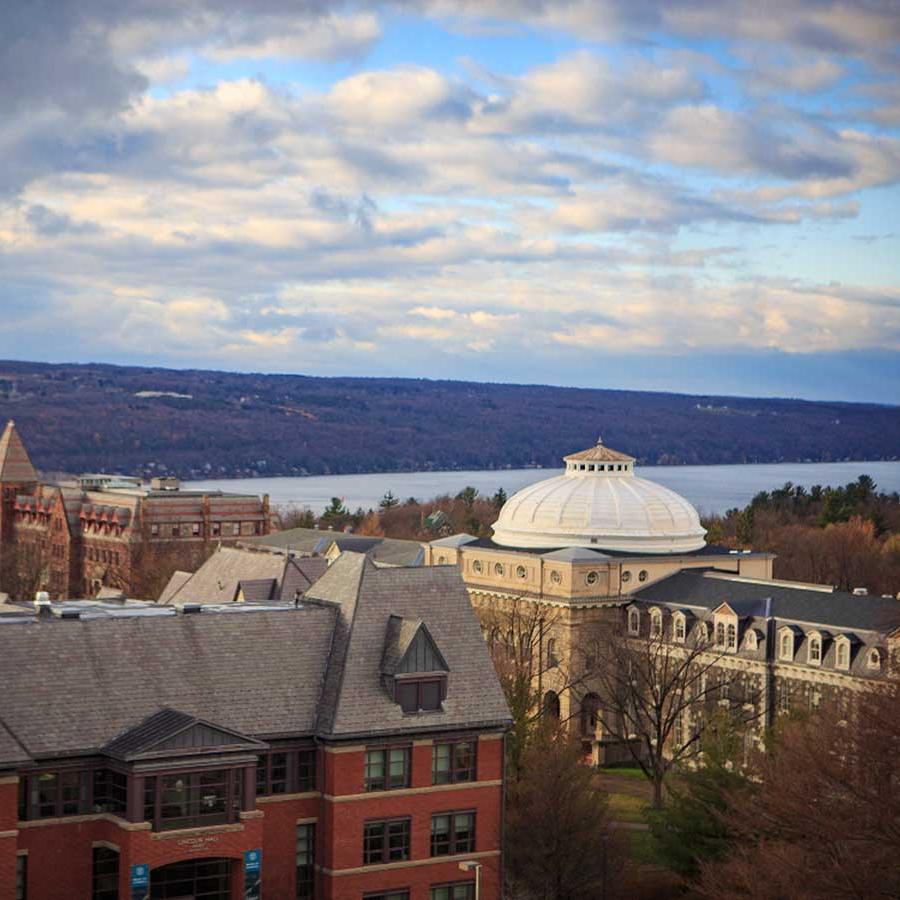 		college campus buildings under a partly cloudy sky, with a lake beyond
	