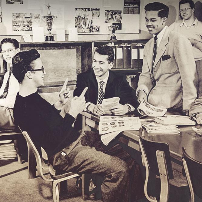 Black and white historic photo of a half dozen young men, relaxing togther around a table