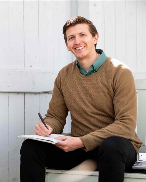 J.J. Zanazzi sitting on a white wooden bench underneath a leafless tree, writing in a notebook; smiling.