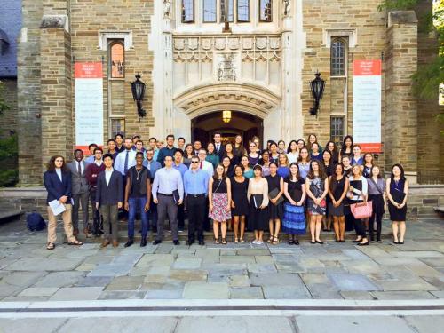 Chorale members standing in four rows in front of a stone building with stone archway