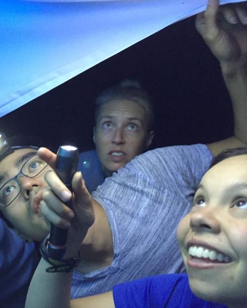 Three people use a flashlight to look at a blue tarp over their heads