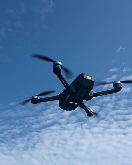 A dark, four-limbed flying drone against a blue sky with fluffy clouds