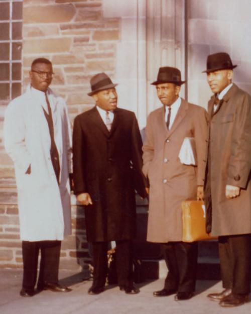 Four people stand in front of a building, wearing dress coats and hats