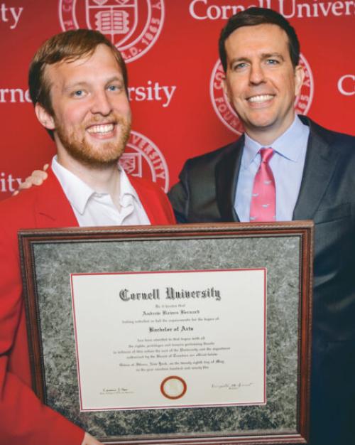 Two people stand in front of a red backgroun, holding a framed diploma