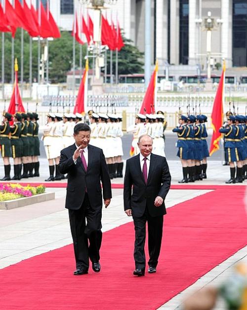 Two people wearing suits walk side by side down a red carpet, waving