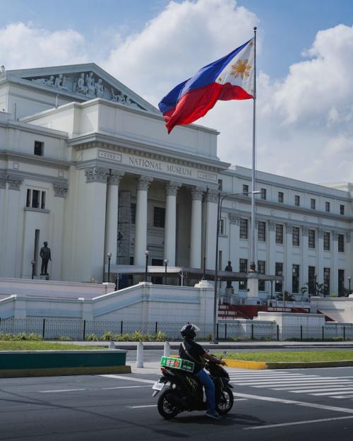 Motorcycle drives past a stone "National Museum" fronted by the Philippine flag