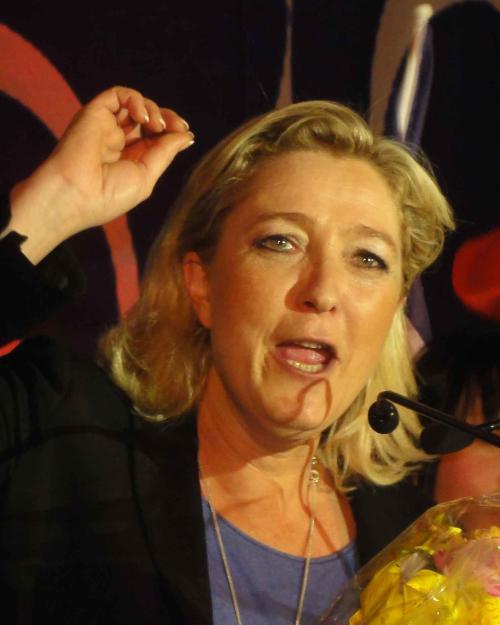 Marine Le Pen with sholuder-length blonde hair and jacket, with hand upraised in the midst of a speech, with French flag in bakcground