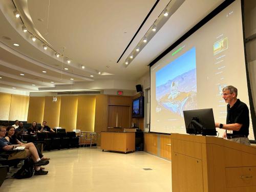 Terry Herter with gray hair and beard standing in front of room at podium with people listening and an image of the Fred Young Submillimeter Telescope on the screen behind him.