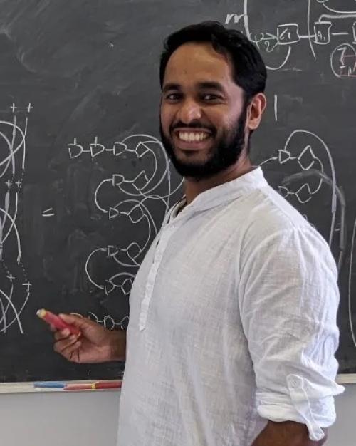 Darren Pereira in a white shirt rolled up to his elbows, smiling with a black beard and mustache, standing at chalkboard in front of diagrams.