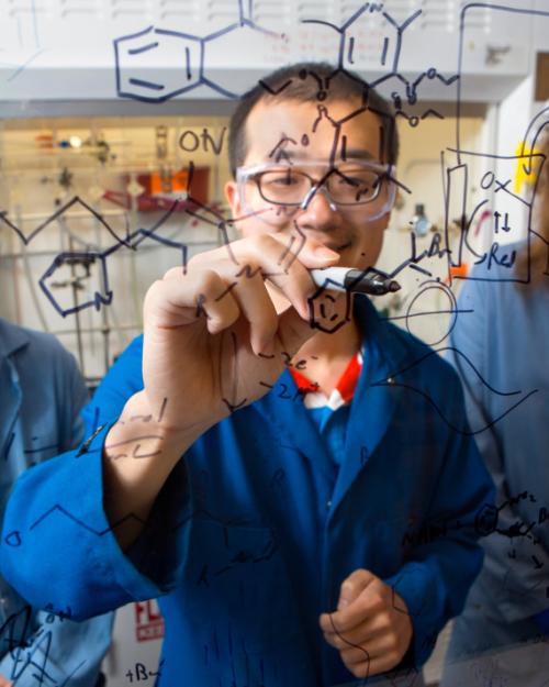 		Three people in lab coats behind a clear wall inscribed with organic chemistry symbols
	