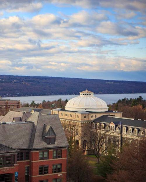 college campus buildings under a partly cloudy sky, with a lake beyond