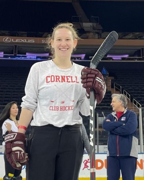 Person wearing hockey gloves and a Cornell t-shirt, carrying a hockey stick and smiling