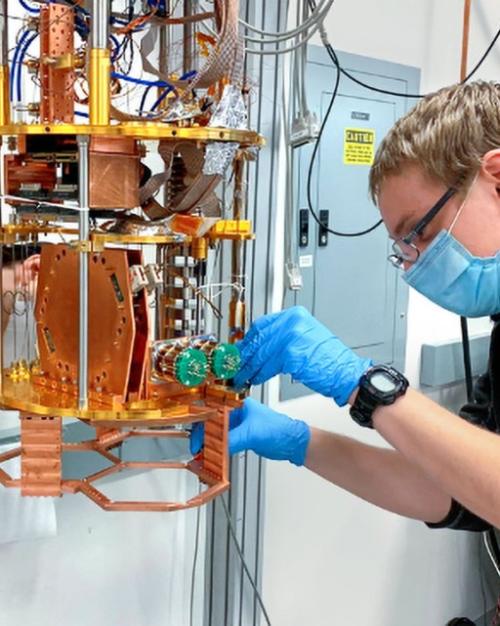 		Two people lean toward a complicated scientific instrument featuring gold and orange metal parts; it's about the size of a coffeemaker
	