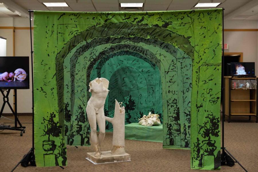 A headless and armless plaster cast with mushrooms growing out of it within a green fabric frame with painted arches 