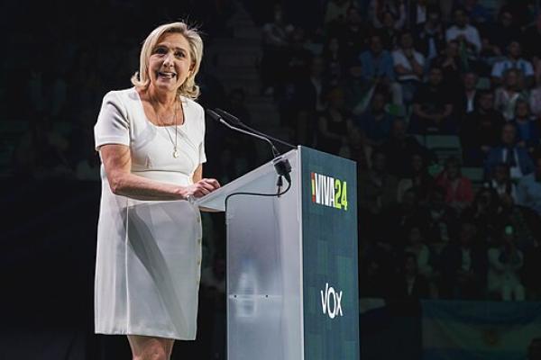 Marine Le Pen in a short white dress facing the audience, standing at a podium that says "Viva24"