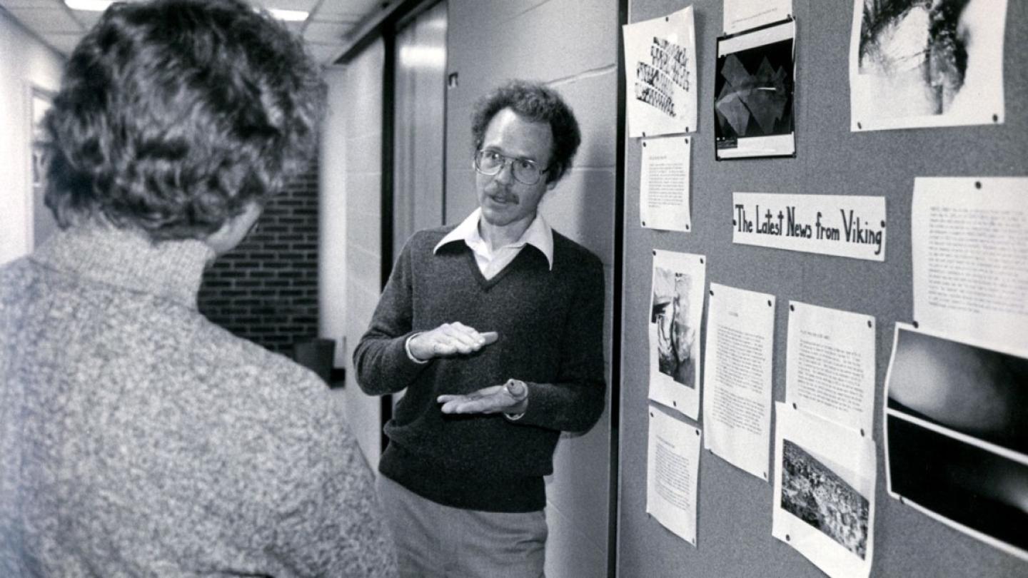 Black and white historic photo: a serious person leans against a wall, explaining something