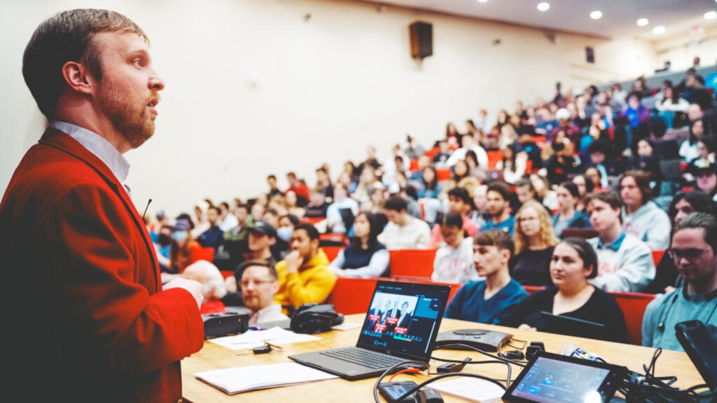 Person in red jacket speaks to a full auditorium