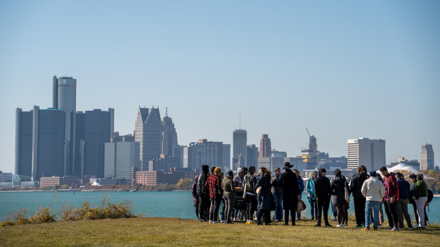 Several people stand on a grassy space looking over a river with a city on the other side