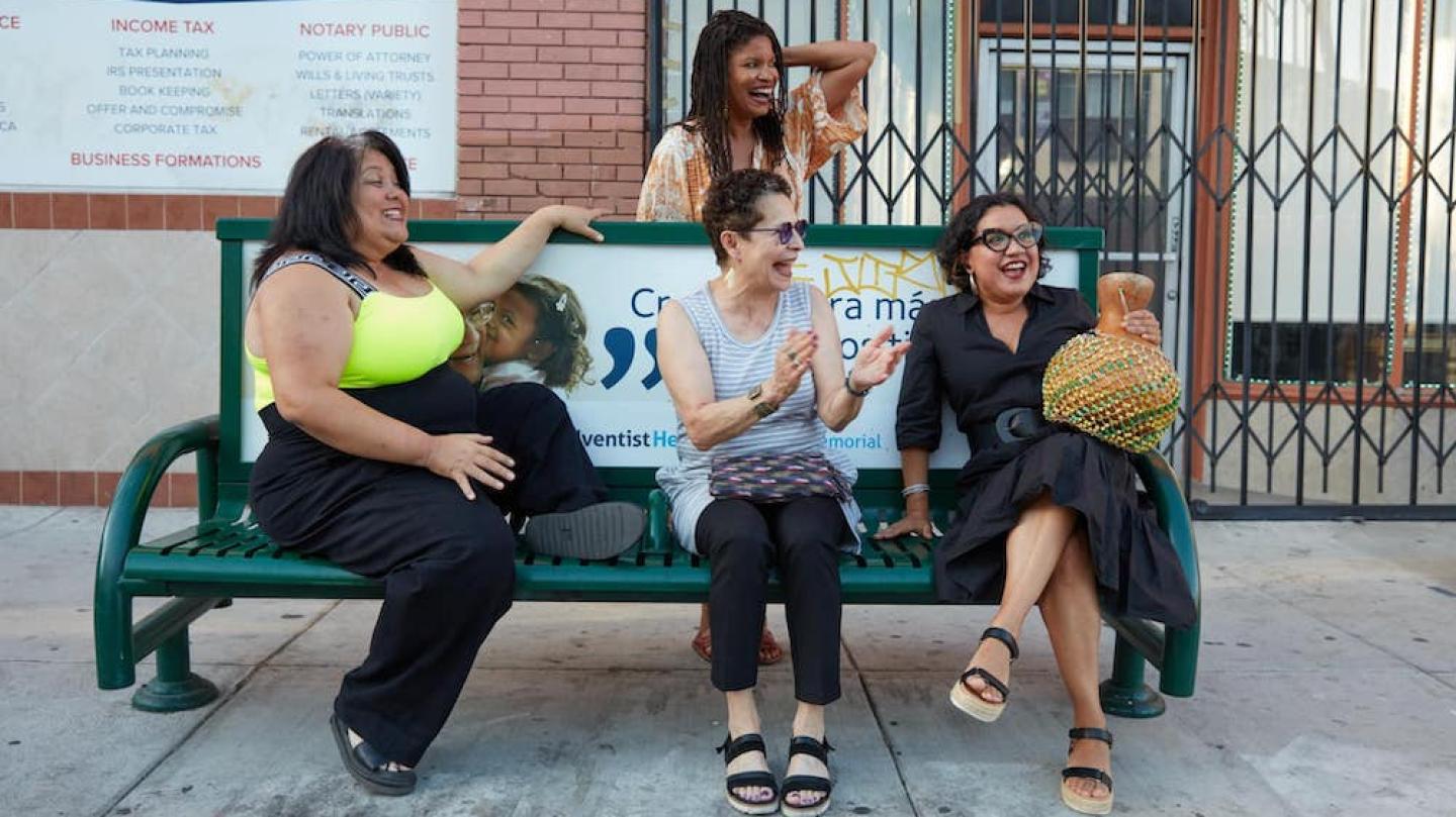 Three people sitting on a city bench with one standing behind; they are laughing together